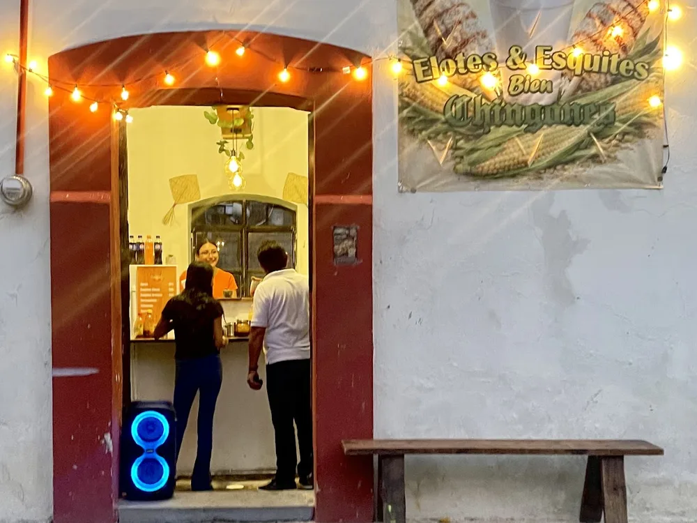 La entrada de El Maiziado al atardecer: puerta con marco guinda, foquitos y la manta de elotes y esquites