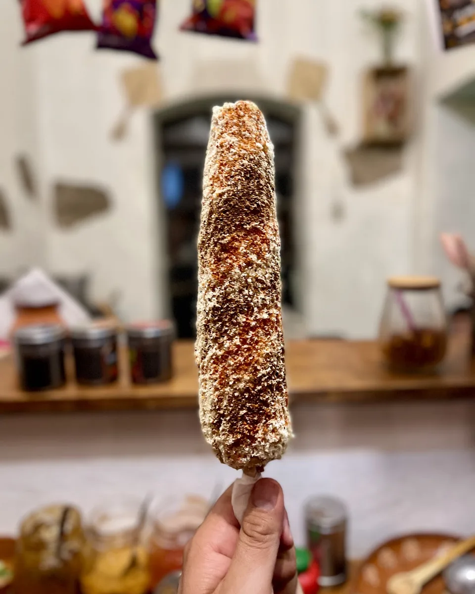 Un elote con mayonesa, queso y chile en la mano, frente a la barra de El Maiziado